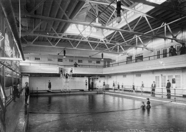 View of a natatorium swimming pool, which opened around 1888. Four people stand in the pool while other people stand around the edge. People are watching by the railing of the balcony near a hallway leading to hot baths.
