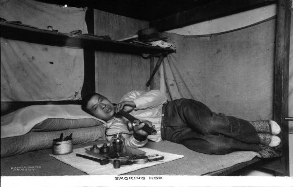 A man in a bunk smokes hop (heroin) from a pipe as he lies on a pillow.  Smoking supplies are arranged on a cloth beside him and a shelf on the wall behind him holds a hat and other belongings. Caption reads: "Smoking Hop."