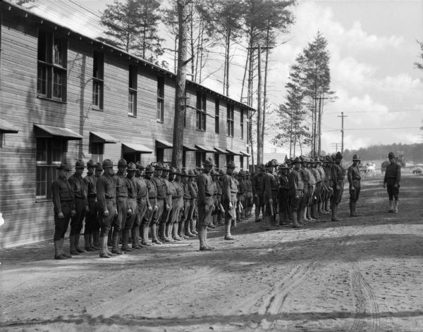View of a group of uniformed men in formation in front of their barracks, awaiting retreat call. The men are lined up with an officer at the front.
