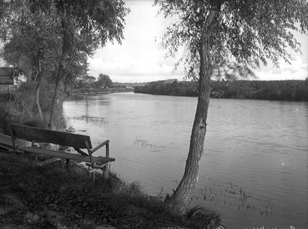 A wooden bench provides a place to look across the Fox River from the left bank. Buildings stand on either side of the river and the opposite shore is heavily lined with trees.