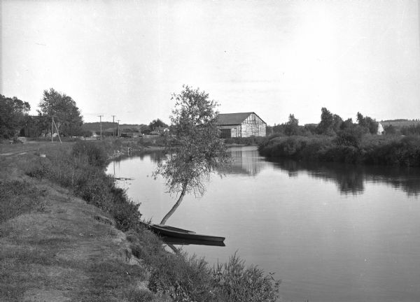 A rowboat floats along the shore of the Fox River near a small tree.    Beyond this, a wooden building stands on the shore and other buildings are visible in the background.