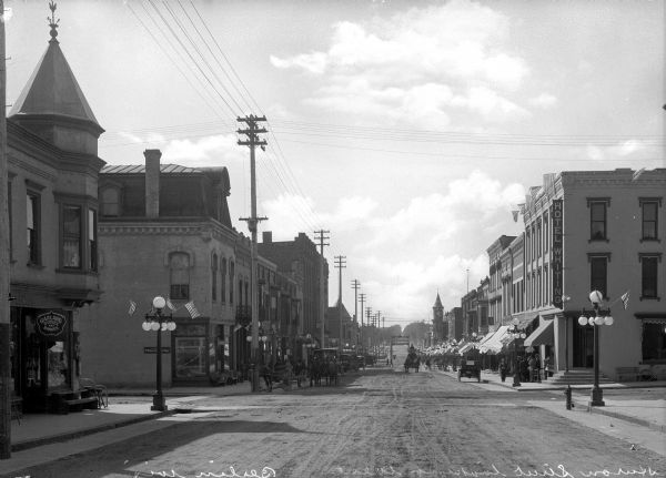 View of Huron Street, looking west. Horse-drawn carriages are parked on either side of the road, and Hotel Whiting can be seen on the right, built in 1868.