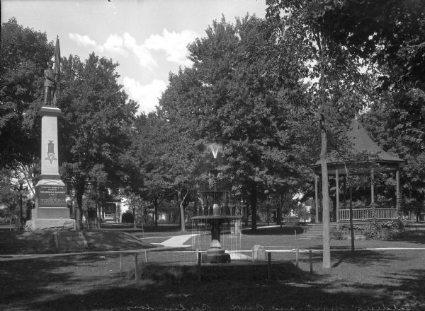View of Nathan Strong Park, a recreation area set aside in 1848. The park features a Volunteer Soldiers and Sailors Monument, topped with a soldier holding a flag. The monument was erected by John H. Williams Post No. 4 C.A.R. and Woman's Relief Corps No. 12. A fountain stands in the middle of the park with a gazebo to its right. A Victorian house built in 1882 stands behind the trees in the background.