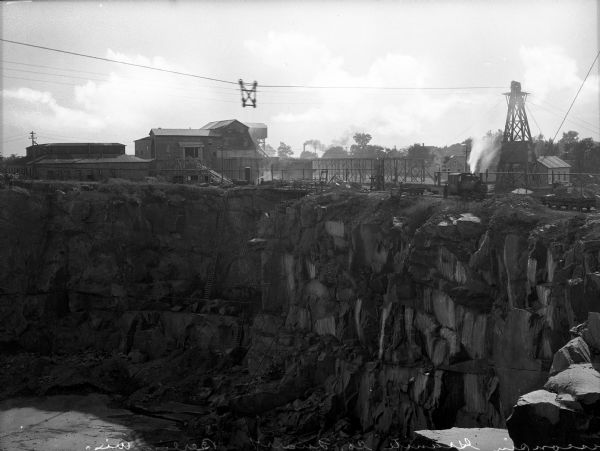 Ladders extend into the quarry at Wisconsin Granite Company, and factory buildings and towers can be seen above. Three men stand near a railroad car at the edge of the quarry. The quarry was founded in 1883 by E.S. Pike and McGinnis.