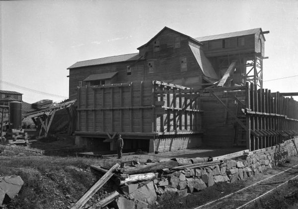 A man stands outside the rock crusher at the Wisconsin Granite Company quarry.  To the right, railroad tracks span the side of the building.