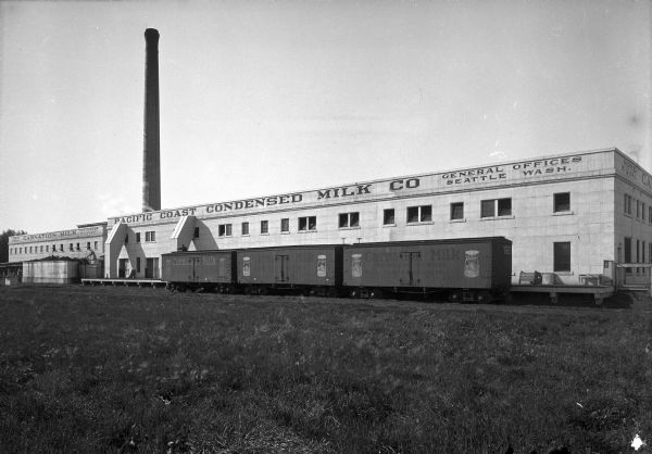 Exterior of Pacific Coast Condensed Milk Company, founded in 1899 by E. B. Stuart. Signs on the factory building read, "None Equal," "The Modern Milk Man," and "General Offices Seattle Wash." Railroad cars labeled "Carnation Milk" are parked in front of the building.