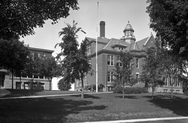 Exterior of High School and Training School. The building features a cupola and brickwork. To the left, a horse-drawn carriage is parked, and a sidewalk leads to another brick building.
