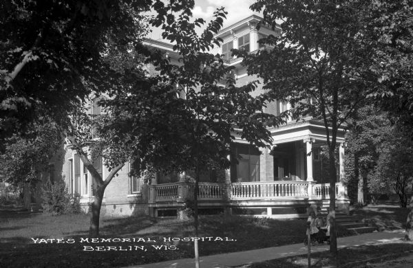 Stairs lead from the sidewalk to a columned porch at the hospital entrance. Three girls play with a wagon on the sidewalk. Caption reads: "Yates Memorial Hospital, Berlin, Wis."