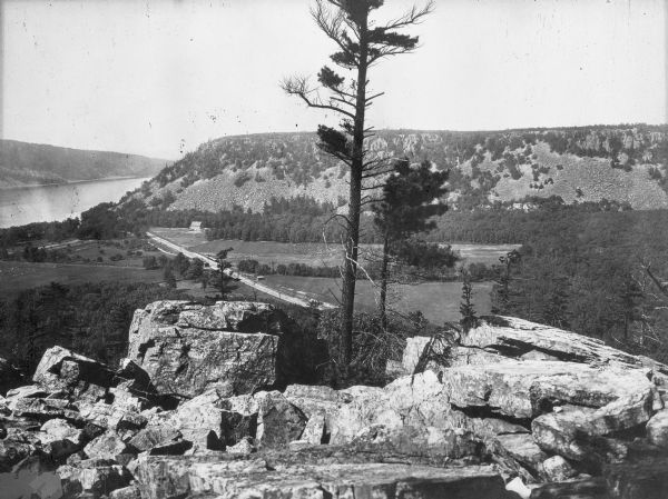 Elevated view from rocks overlooking a valley toward Devil's Lake. Several houses are near a road that leads to the shoreline.