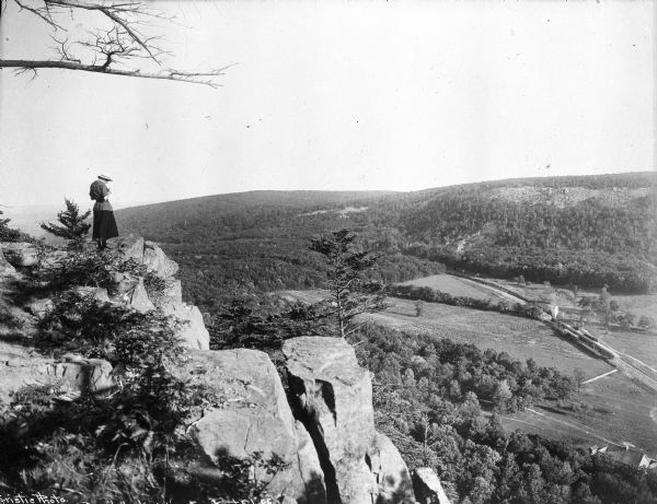 View of a woman taking a photograph from Lookout Cliff at Devil's Lake. Below, a train runs through a field surrounded by trees.