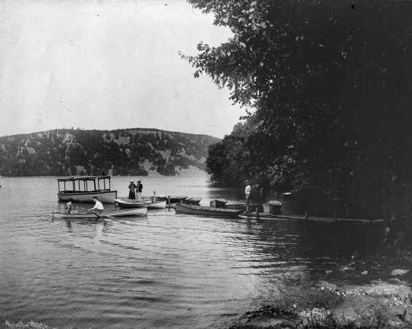 A man and two women stand on a dock at Devil's Lake while a couple rows in a canoe on the water.  Other rowboats are docked nearby and a covered rowboat named "Texas" floats at the end of the dock.