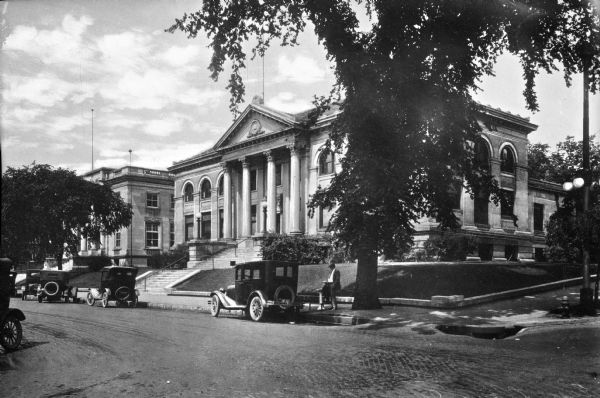 A view from across the street shows the staircase leading to the main entrance of Eau Claire Public Library. A man and child stand amidst automobiles parked along the side of the street. The Eau Claire Public Library opened in 1904 after a 1902 Carnegie Grant.