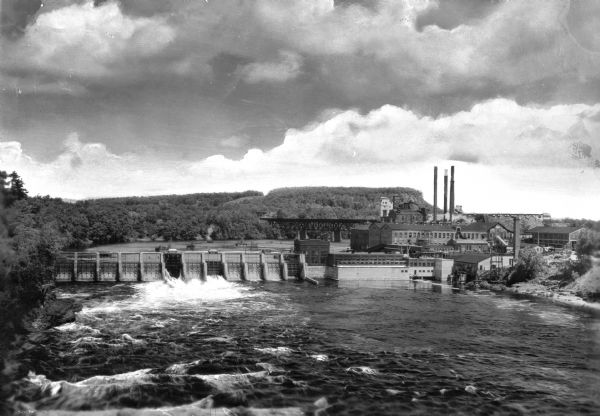 Elevated view looking upriver toward the Dells Paper and Pulp Company factory which stands near a dam on the Chippewa River. A bridge, smokestacks, and several other buildings are nearby. The Dells Paper and Pulp Company opened in 1890.