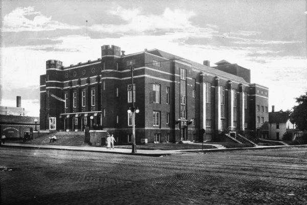 View across intersection toward a brick auditorium building. A man sits on the steps of the main entrance while others walk on the sidewalk. Three men in costume sit on the side wall of the steps. In the background is a Northern Pacific train.