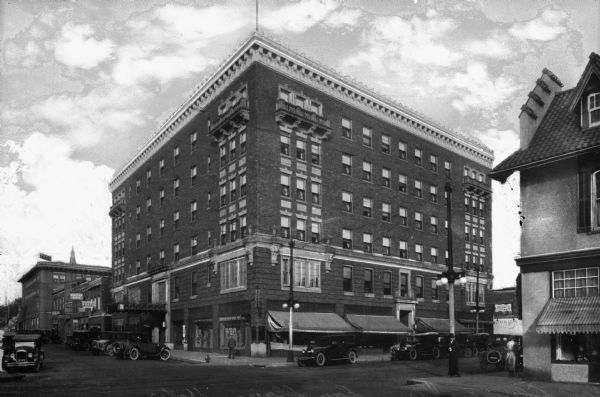 Exterior view from street of the Eau Claire Hotel, located on the corner of South Barstow and Eau Claire Street. Automobiles are parked along the curbs, and pedestrians are on the sidewalk along the storefronts. On the left is the YMCA, and signs read: "Western Union," "Furs," and "Photo Service." A coffee shop is on the right.