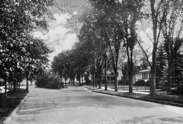 View down a residential street, partially divided by a median planted with trees and shrubs. On the right fences separate the sidewalk from the front lawns of houses.