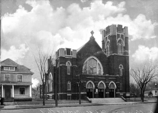 Front view of Methodist Church located in a residential area on Lake Street. Stairs lead to the main entrance that features three doors topped by Gothic arches. Above, a large pointed-arch window can be seen below a cross on the roof. A belfry rises on the right.