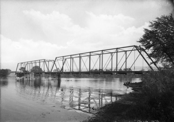 View from shoreline toward an unidentified railroad bridge spanning a river.