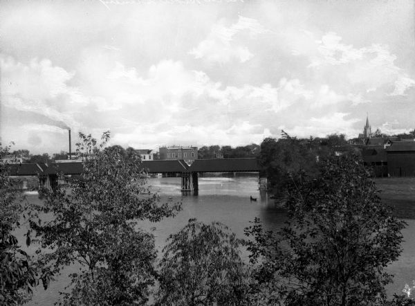 View over tree-tops toward a man rowing a boat beneath a partially covered bridge spanning the Rock River. Factories, industrial buildings, and Saint Paul's Lutheran Church are on the opposite riverbank.