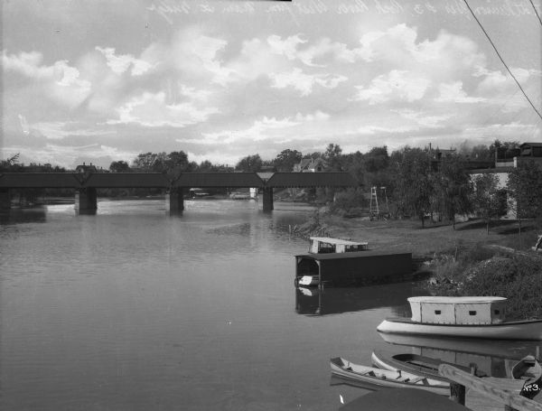 Elevated view looking west from the Main Street Bridge, a partially covered wooden bridge spanning the Rock River. In the foreground on the right are boats dock along the shore. Riverside dwellings are in the background.
