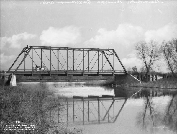 View from the left side riverbank of the Bark River, whose water is reflecting the bridge above. A man is driving an automobile over the bridge toward the right bank.