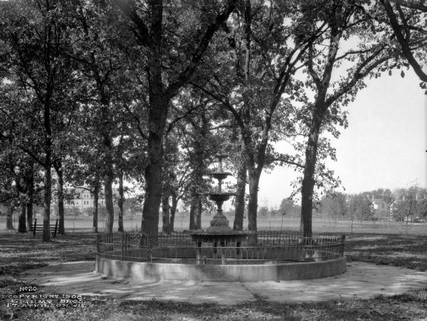 View of Jones Fountain at Barrie Park, erected in 1905. The fountain stands among trees and a park bench, and dwellings are in the background.