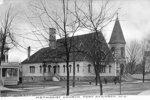 Exterior view across road toward the Methodist Church. Children play with a wagon on the sidewalk in front of the church. Two sets of stairs lead to a side entrance and to a main entrance beneath a bell tower. Caption reads: "Methodist Church, Fort Atkinson, Wis."