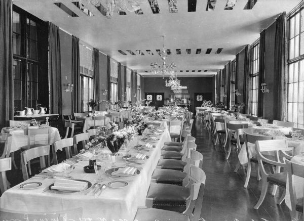 Interior view of Lawsonia Country Club, which opened to the public in 1930. The dining room features long and circular tables set for a meal. Chandeliers and mirrors are affixed to the ceiling.