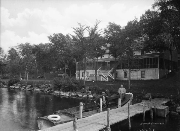 Rear view of the Bible Institute on Green Lake. Two men are sitting in a boat docked at a pier. Behind them are stairs leading to the rear entrance of the Bible Institute on the shoreline.