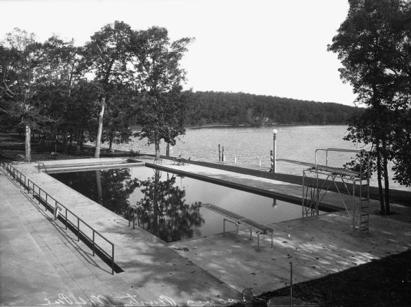 View of a swimming pool at Lawsonia Country Club, which opened to the public in 1930. Two diving boards are in the foreground at one end of the pool. Tables and chairs are placed around the pool, which is overlooking Green Lake.