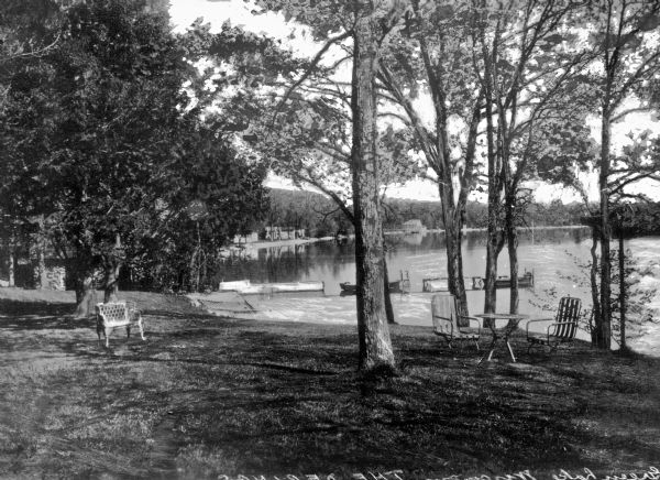 View of Dering's yard on Green Lake. The yard features outdoor furniture on the lawn among trees, and boats are docked along a pier behind them.