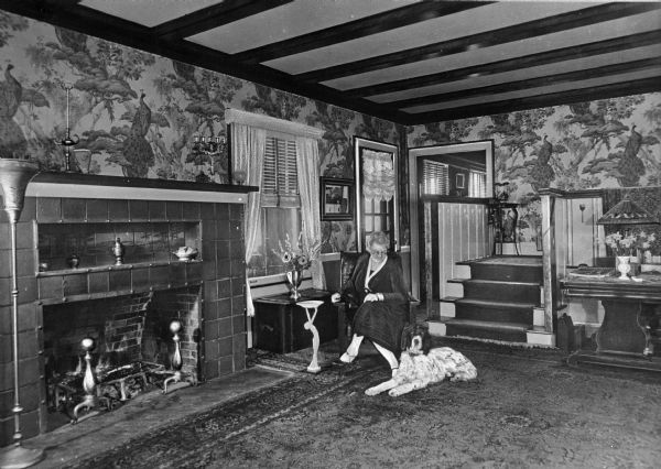 Interior of Dering's living room. A woman sits in a chair looking at a dog that is lying on the rug. A fireplace is on the left, and the walls are lined with decorative peacock wallpaper. In the background is a set of stairs leads to a walkway.