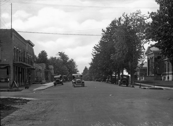 View down Hill Street. An automobile is crossing an intersection while other automobiles are parked along the curbs. The Green Lake County Courthouse, built in 1863, stands to the right. A boy plays in a wagon and two young girls walk past storefronts on the left sidewalk.