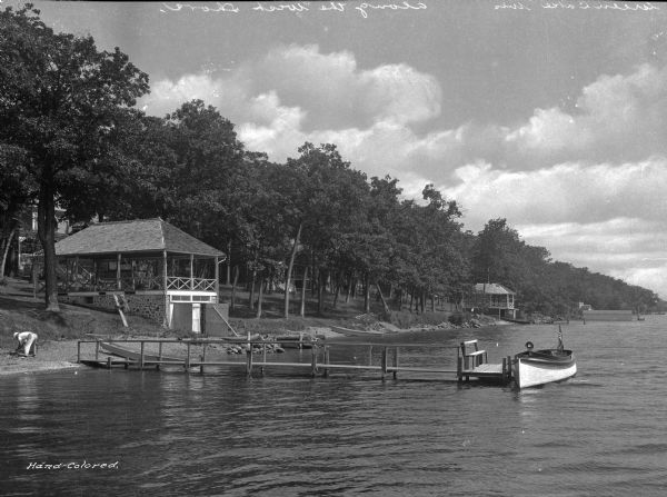 View along the west shore of Green Lake. Boats are docked on the shore near boathouses and two men are on the left, one carrying a piece of wood.