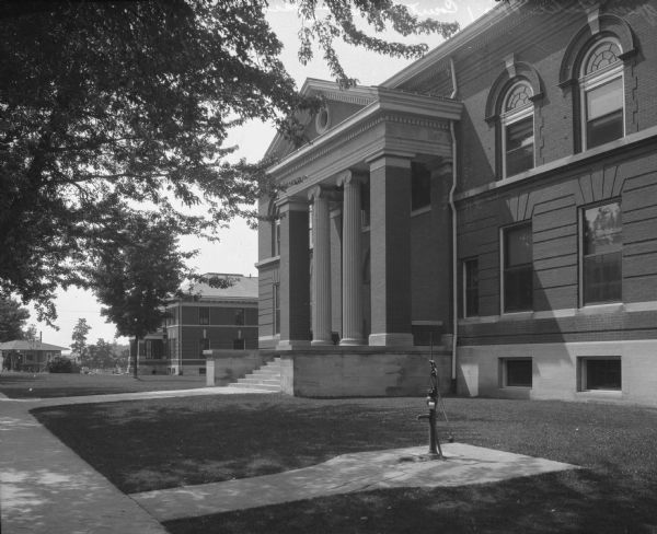 View of the Green Lake County Courthouse and Jail. The courthouse, built in 1899, features stairs leading to a central pavilion. The jail can be seen to the left. In the foreground is a hand-pump.