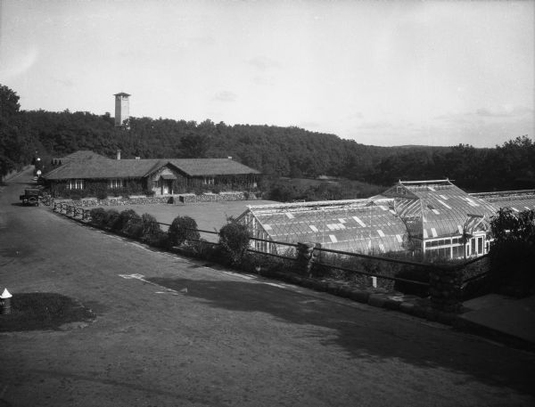 View of a greenhouse. An ivy-covered building and a watch tower are visible to the left of the greenhouse. An automobile is parked on a street, separated from the buildings by a railing.