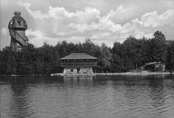 View across water towards the Mayor's summer home as seen from across Pine Lake. Children and adults are standing in front of the two-story dwelling. To the building's right are stairs leading to a road in the woods. A domed lighthouse stands high above the trees on the left.