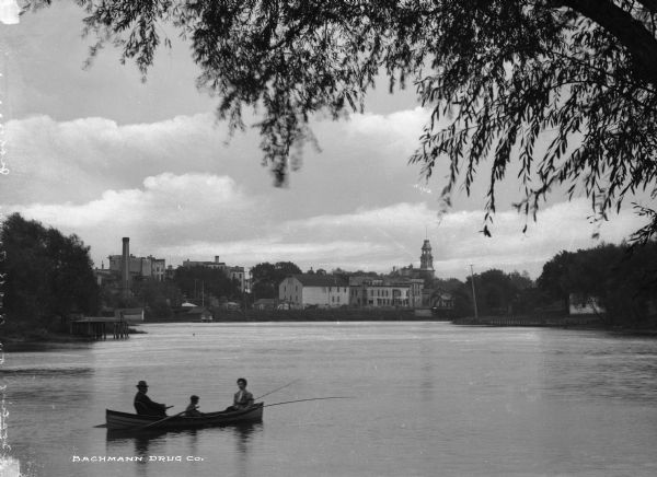 View of a man, woman, and child boating on the Rock River. A church spire and factory buildings are on the opposite shore in the background.