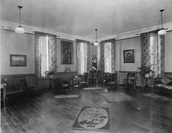 Interior of the reception room at Saint Clara Academy. Founded by Father Samuel Mazzuchelli and built in 1852, the academy's reception room features religious sculptures and paintings. Decorative curtains and rugs adorn the room near chairs, tables, lamps, and potted plants.