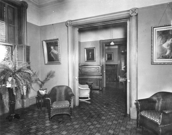 Interior of reception rooms at Saint Clara Academy. Founded by Father Samuel Mazzuchelli and built in 1852, the academy's reception rooms feature religious paintings, potted plants and wicker chairs. Through pocket doors, two pianos can be seen in separate rooms.