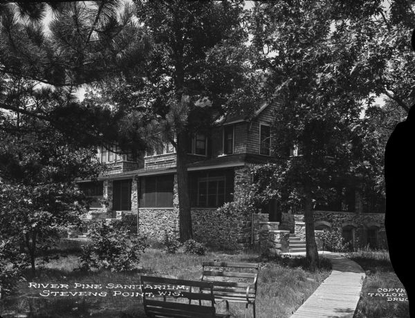 Exterior of River Pine Sanitarium, which opened in 1906. A path surrounded by trees leads past benches to the sanitarium porch. Caption reads: "River Pine Sanitarium, Stevens Point, Wis."