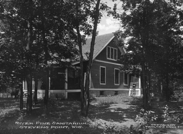 Exterior of River Pine Sanitarium, which opened in 1906. Trees surround a sanitarium building with porches on the front and back. Caption reads: "River Pine Sanitarium, Stevens Point, Wis."  