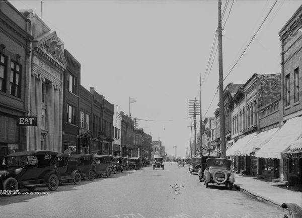 View down Main Street. Cars are parked on either side of the street, and two cars are being driven by storefronts. On the left stands the Citizens National Bank, built in 1893.
