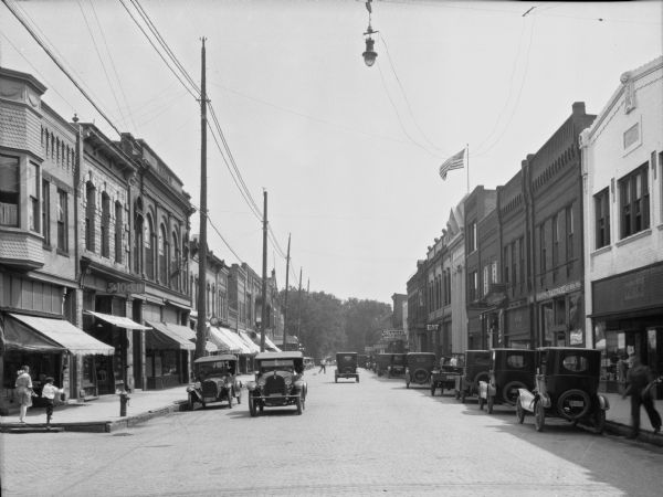 View down Main Street. Cars are parked on either side of the street and two cars are being driven by storefronts. Citizens National Bank, built in 1893, stands to the right.