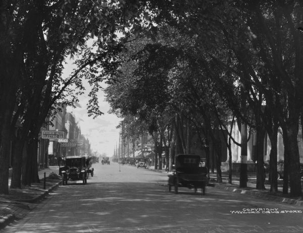 View down East Main Street lined by trees arching over the street. Cars are parked along the curbs, and two cars aredriving by storefronts.