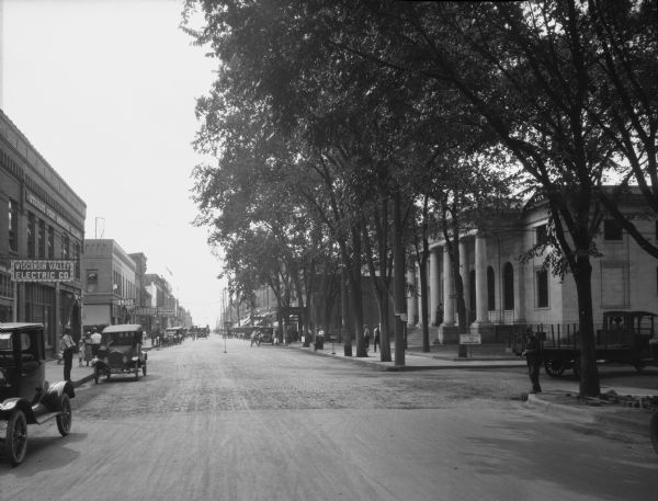 View down street of business district with automobiles parked along the curbs. Pedestrians are on the sidewalks. Trees are along the right side of the street. The Wisconsin Valley Electric Company stands on the left; the company merged with the Stevens Point Power Company in 1916.