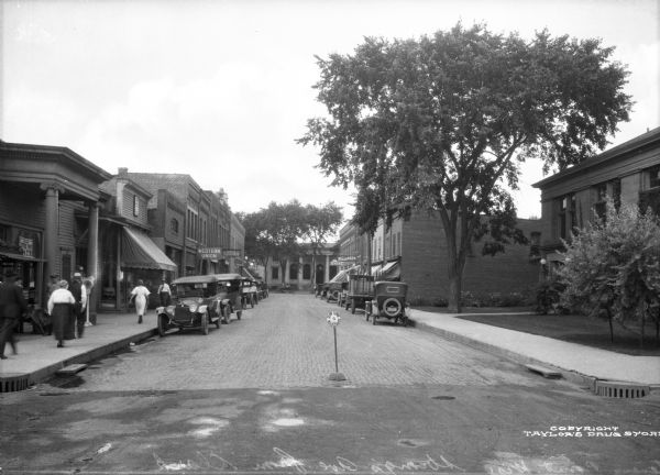View down Strong's Avenue, taken from Clark Street. Automobiles park on either side of the street near storefronts. Perpendicular to Strongs Avenue is Main Street, featuring the city's post office, a classical revival structure built in 1913. A sign near the intersection in the middle of Strongs Avenue reads: "Slow Down & Keep Right." Pedestrians can be seen walking past stores and businesses