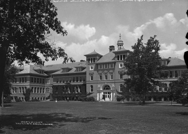 Exterior of the State Normal School at Stevens Point, which opened in 1894. The main entrance is flanked by columns, a central cupola, and a row of pedimented windows. Two girls sit outside the entrance.