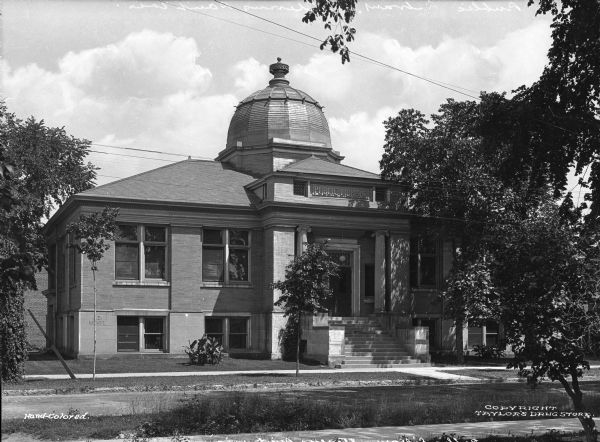 Exterior of the Carnegie Public Library, built in 1903 after the receipt of a 1902 Carnegie Grant. A set of stairs leads from the sidewalk to the main entrance where two Ionic columns can be seen on either side of the double doors. The library building is topped by a segmented dome.