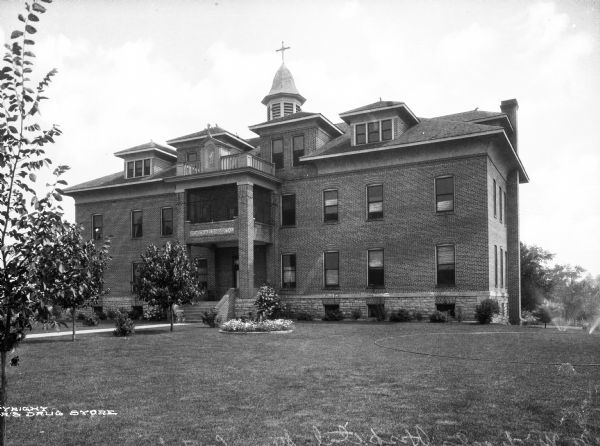 Exterior of Saint Michael's Hospital, founded in 1913 by the Sisters of the Sorrowful Mother. A baby carriage stands near the bottom of the steps at the hospital's main entrance and a man sits on the porch of the brick building. A niche houses a religious statue on the top balcony.
